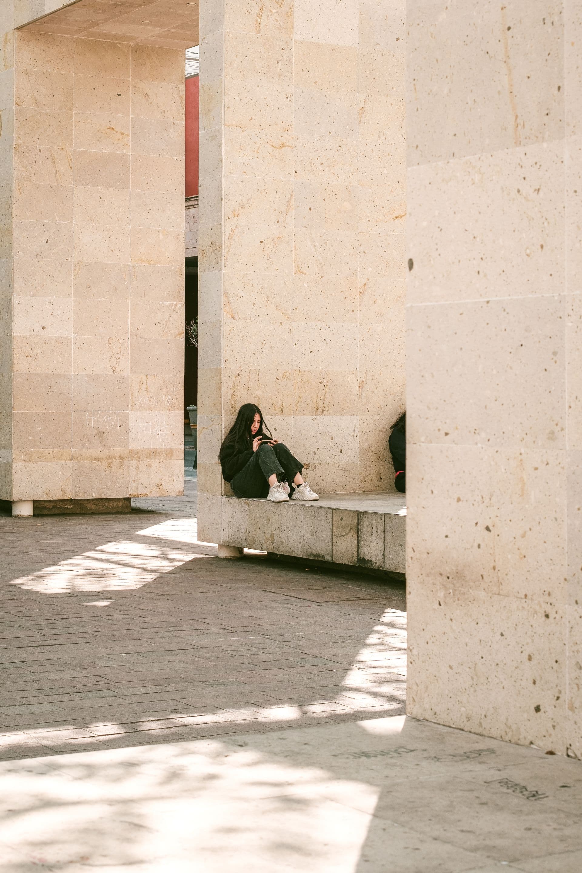 A person sitting alone on a stone bench in a sunlit courtyard, small against tall limestone walls.